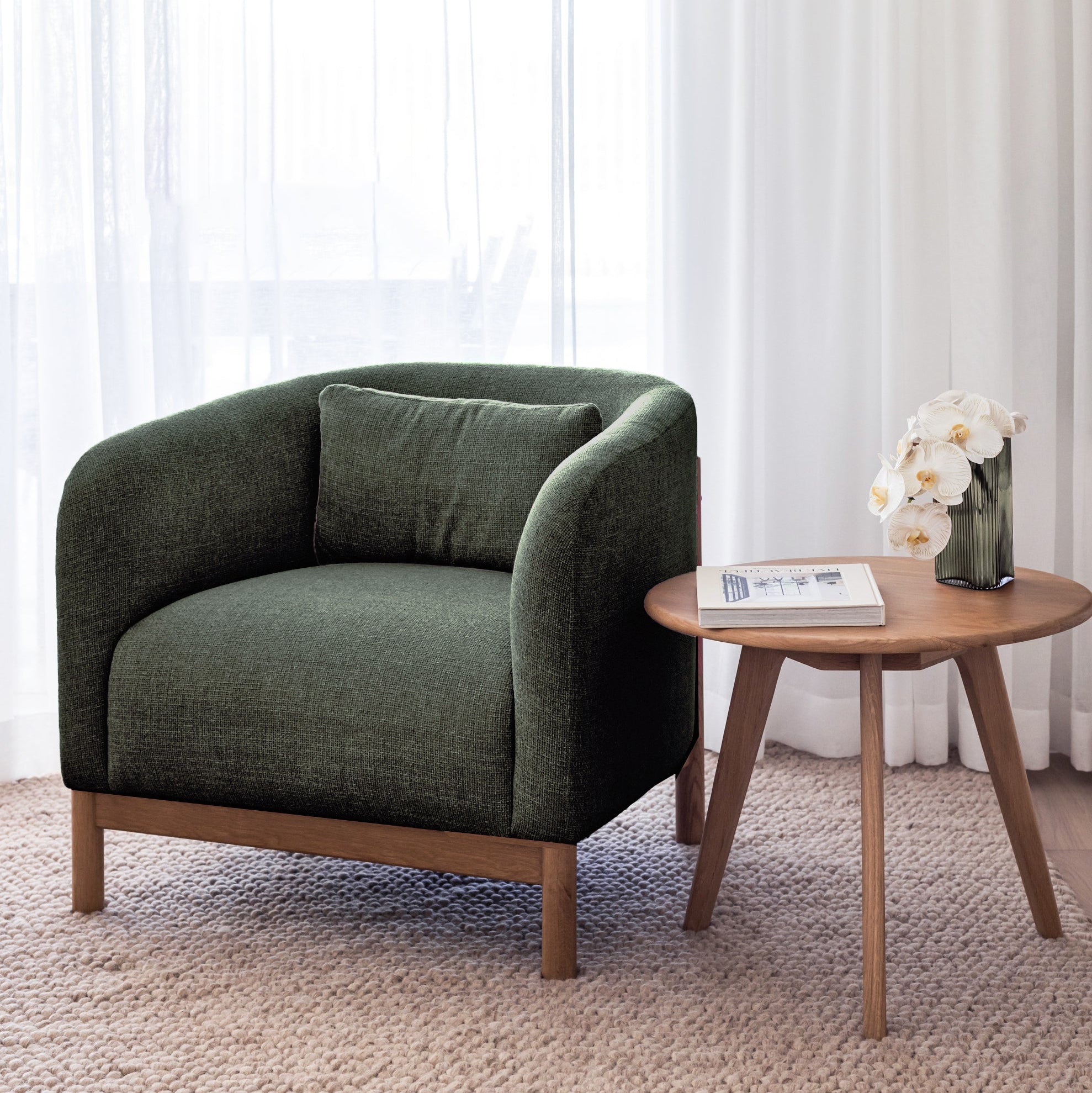 Green armchair with wooden legs in a room with white curtains and a small table with a vase of flowers.